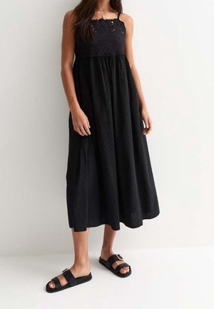 Žena v černých midi šatech bez rukávů s krajkovým vrškem a volánovou sukní, doplněná černými sandály se sponou, stojící na jednobarevném pozadí.