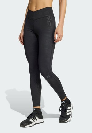 Kobieta ubrana w czarne legginsy w wzory i czarno-białe buty sportowe Adidas, stojąca na tle jednolitego jasnego tła.