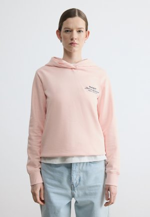 Jeune femme portant un sweat à capuche rose clair avec un texte sur la poitrine et un jean bleu clair, debout devant un fond blanc uni.