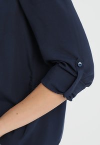 Marineblaue Bluse mit weicher Textur, mit aufgerolltem Ärmel, der durch einen passenden Knopf befestigt ist, und einem locker geschnittenen Design.