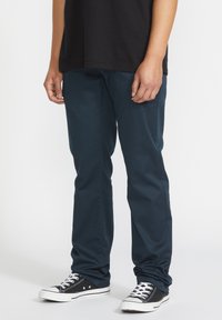 Dark blue trousers made of a smooth fabric, featuring a slim fit design. Paired with black high-top sneakers with white accents.