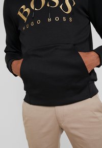 Svart kapuschongtröja med guld "BOSS HUGO BOSS" logotyp, framficka, och händerna i fickan, ihopparad med beige byxor.