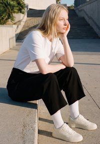 Young woman with long blonde hair sitting on outdoor steps, resting chin on hand, wearing white shirt, black pants, white socks, and sneakers.