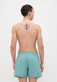 Turkoosit uimashortsit, joissa on joustava vyötärö, vasemmassa lahkeessa logo ja koristeellinen fleur-de-lis -tatuointi takana.