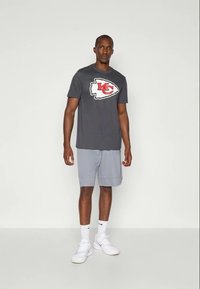 Mand stående iført en mørkegrå Kansas City Chiefs t-shirt, lysegrå shorts, hvide Nike sokker og hvide sneakers på en ensfarvet baggrund.