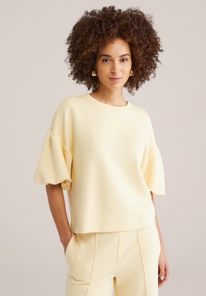 Femme aux cheveux bouclés portant un haut crème à manches courtes bouffantes et un pantalon assorti, debout avec une main dans la poche.