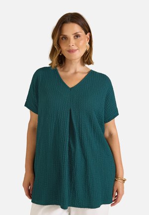 Femme aux cheveux bruns courts portant un haut turquoise texturé à manches courtes et col en V, avec des boucles d'oreilles créoles dorées, sur un fond clair uni.
