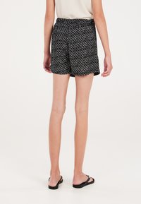Pantaloni corti neri a fantasia con cintura elastica, caratterizzati da un motivo a rombi e una vestibilità ampia. Abbinati a infradito nere.