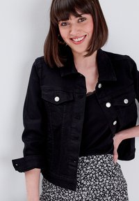 Chaqueta de denim negra con botones plateados y un cuello, con un diseño ajustado y mangas de tres cuartos. Llevada sobre una blusa negra y una falda con estampado.