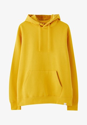 Persona con sudadera amarilla con capucha, pantalones cortos vaqueros azules y zapatillas blancas, de pie frente a un fondo liso y claro.