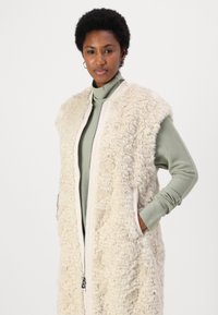 Gilet fluffy color crema con zip frontale, tasche laterali e colletto liscio. Indossato sopra una maglietta a maniche lunghe verde menta.
