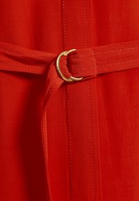 Tissu rouge à texture lisse, doté d'une ceinture maintenue par une boucle circulaire dorée. Comprend des détails de couture subtils le long des bords.