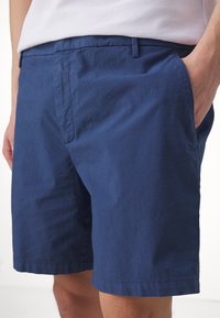 Homme portant un short décontracté bleu marine et une chemise blanche, avec une main dans la poche, montré de la taille à mi-cuisse.