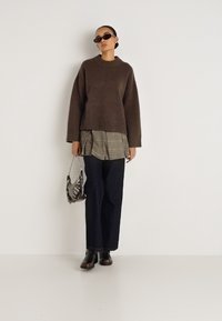 Καφέ πλεκτό πουλόβερ με χαμηλωμένους ώμους, φορεμένο πάνω από πουκάμισο καρό, συνδυασμένο με σκούρο τζιν φαρδιάς γραμμής και μποτάκια μέχρι τον αστράγαλο. Κρατώντας μια τσάντα με σχέδια.