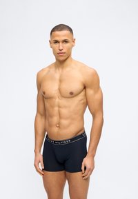 Μυώδης άντρας με κοντά μαλλιά που φοράει μαύρα μπόξερ Tommy Hilfiger, στέκεται μπροστά από ένα απλό ανοιχτογκρι φόντο.