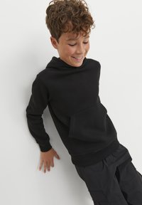 Felpa nera con cappuccio realizzata in morbido tessuto, presenta una tasca kangaroo frontale, polsini a costine e una vestibilità attillata. Indossata con pantaloni cargo scuri.