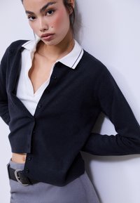 Cardigan bleu marine en tissu doux, doté d'un col en V, de boutons noirs et d'un ourlet côtelé. Superposé à une chemise blanche à col.