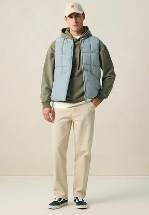 Jeune homme portant une casquette beige, une veste matelassée bleue, un sweat à capuche vert olive, un pantalon crème et des baskets vert foncé, debout devant un fond clair uni.