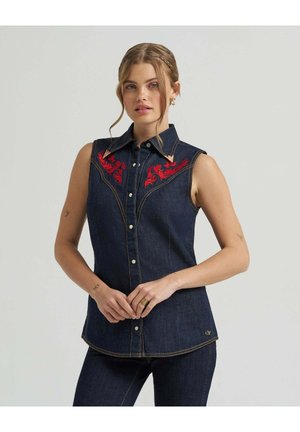 Frau trägt ärmelloses dunkles Jeanshemd mit roten bestickten Motiven auf der Brust und dunkle Jeanshose, steht mit verschränkten Händen.