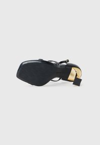 Sandal de salto alto preto com sola texturizada, detalhe metálico dourado no salto e tiras finas pretas. Etiqueta presa ao salto.