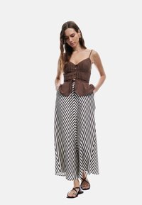 Bruine peplumtop met verticale plooien en knoopdetails, gecombineerd met een lange zwart-wit gestreepte rok. Model draagt strappy zwarte sandalen.