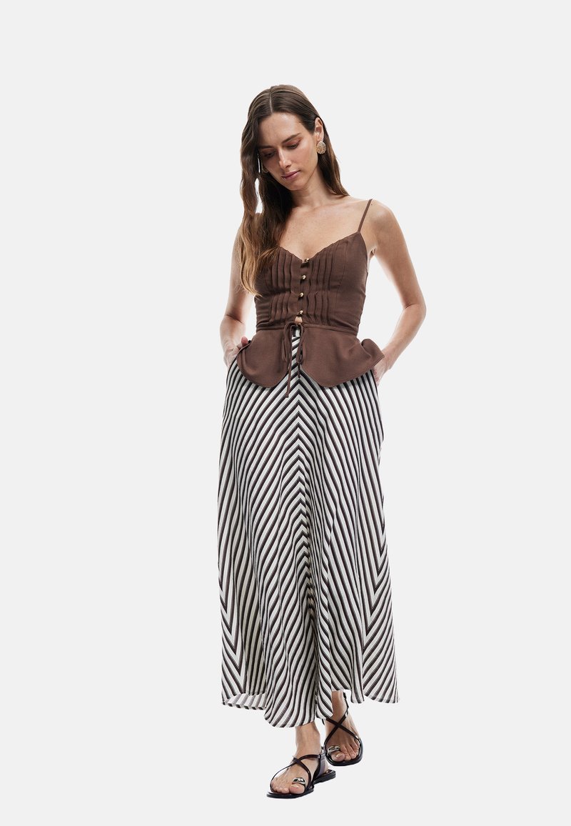 Bruine peplumtop met verticale plooien en knoopdetails, gecombineerd met een lange zwart-wit gestreepte rok. Model draagt strappy zwarte sandalen.