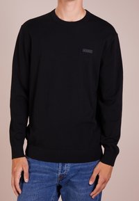 Hombre vestido con un suéter negro de manga larga con un pequeño parche de logo negro en el pecho, combinado con jeans de mezclilla azules, frente a un fondo neutro.