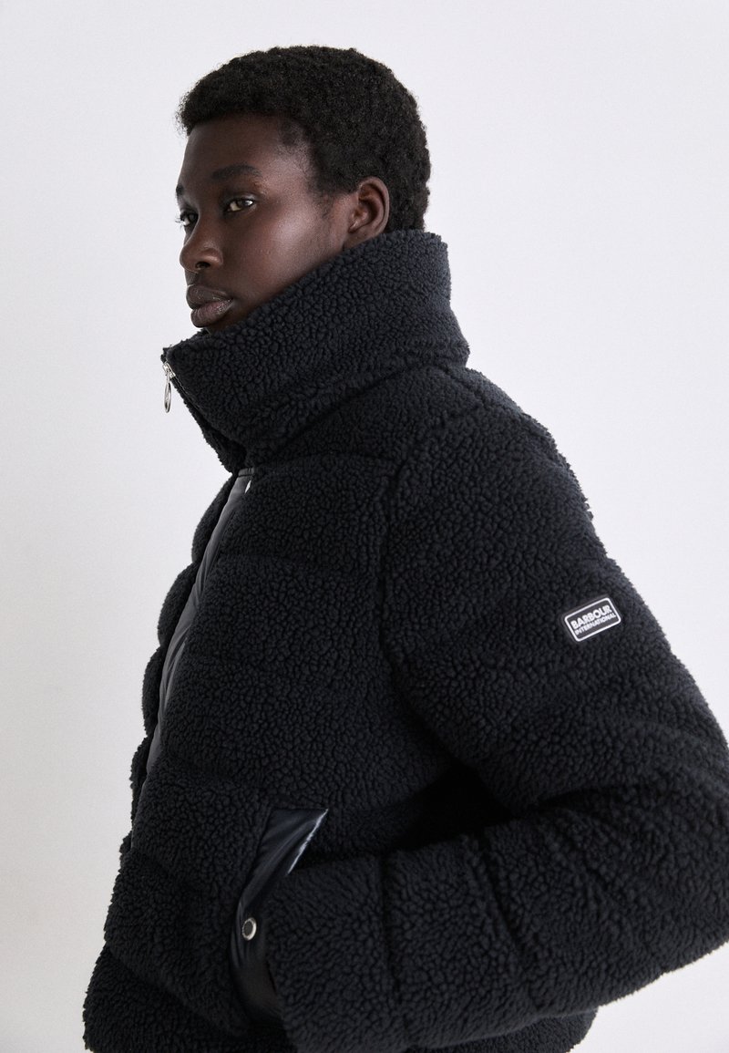 Giubbotto imbottito in faux fur nero con collo alto, zip laterale e pannello liscio a contrasto. Presenta una toppa con logo sulla manica.