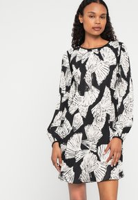 Rakstainā kleita ar melnbaltu čaulas dizainu, garām piedurknēm, kroku tekstūru un apaļu izgriezumu. Īsais garums.