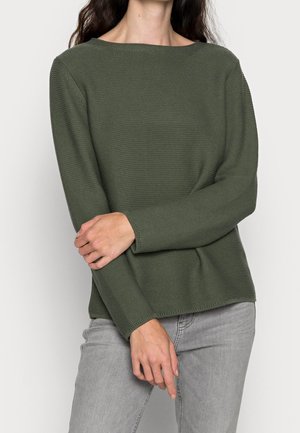 Vrouw die een olijfgroen gebreide trui met textuur en lange mouwen draagt, gecombineerd met lichtgrijze spijkerbroek.