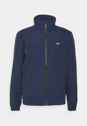 Navy blue zip-up jacket with high collar, two front pockets, elastic cuffs, and small rectangular logo on chest and left sleeve.