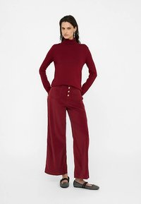 Pull à col roulé bordeaux et pantalon large assorti avec trois boutons sur les poches avant. Le mannequin porte des ballerines noires à finition texturée.