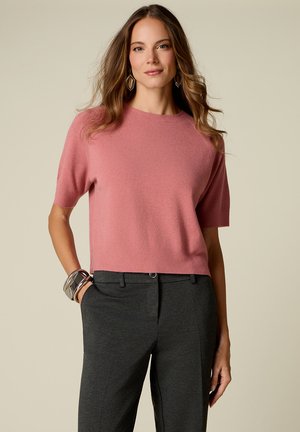 Vrouw met golvend bruin haar die een roze sweater met korte mouwen, donkergrijze broek, geometrische oorbellen en zilveren armbanden draagt.