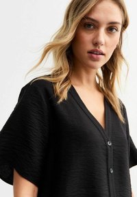 Zwarte top met korte mouwen heeft een V-hals en knoopdetail. De stof heeft een gestructureerde afwerking, wat zorgt voor een zachte, casual uitstraling.