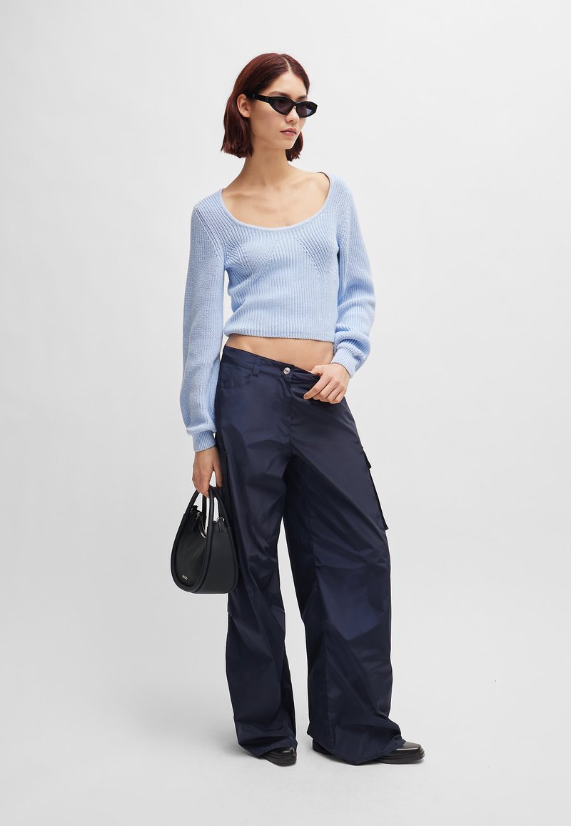Hellblaues gestricktes Crop-Sweatshirt mit langen Ärmeln; kombiniert mit navyfarbenen, weiten Cargo-Hosen. Das Model hält eine schwarze Handtasche.