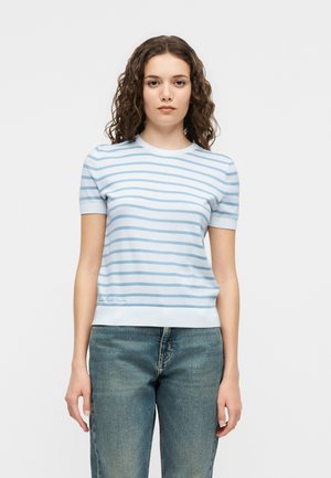 Mladá žena s kučeravými vlasmi, ktorá má na sebe biele krátke tričko s horizontálnymi svetlomodrými pruhmi a modré džínsy, stojaca na jednoduchom pozadí.