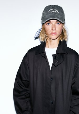 Personne blonde aux cheveux courts portant un manteau foncé, une chemise blanche et une casquette grise "Saint Louis Paris" sur un fond blanc.