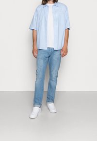 Man som bär en ljusblå randig kortärmad skjorta över en vit t-shirt, ljusblå jeans och vita sneakers, står mot en neutral bakgrund.