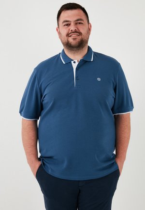 Plava polo majica s bijelim ovratnikom i naglascima na rukavima, sa dugmadima, teksturirana tkanina, nošena s tamnoplavim hlačama, sa logotipom na prsima.