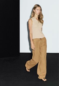 Woman walking against black and white background wearing beige sleeveless top, loose brown pants, and black flip-flops.