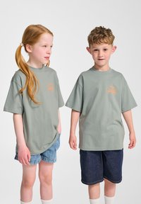 Gröna bomullströjor med kort ärm, med en liten orange logotyp på framsidan, i kombination med denimshorts.