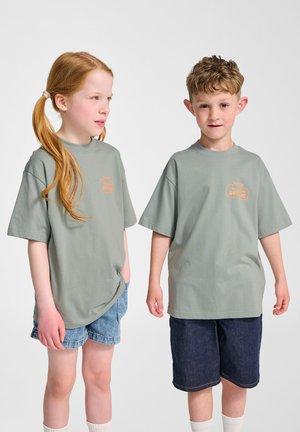 Grüne Baumwoll-T-Shirts mit kurzen Ärmeln, die ein kleines oranges Logo auf der Vorderseite tragen, kombiniert mit Jeansshorts.