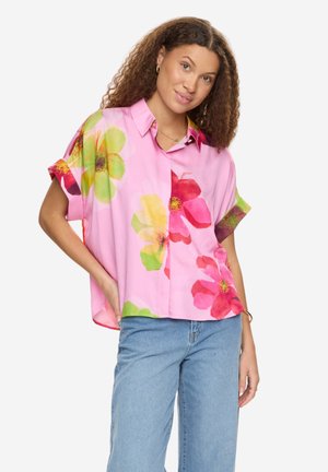 Kobieta o kręconych włosach, ubrana w różową bluzkę z krótkim rękawem z dużymi, wielokolorowymi kwiatami i jasnoniebieskie dżinsy, stojąca z jedną ręką w kieszeni.