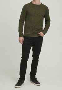 Man in een olijfgroen gemêleerd sweater, zwarte broek en zwarte sneakers, staand met één hand in de zak tegen een effen achtergrond.