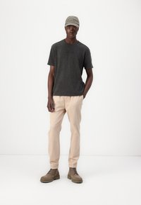 T-shirt gris avec logo noir, pantalon beige et bottes à lacets marron. La casquette est gris clair avec du texte. Tenue simple et décontractée.