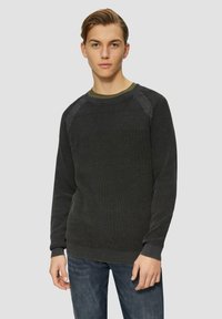Dunkelgrüner Strickpullover mit strukturiertem Muster und gerippten Bündchen. Über einem hellgrünen Hemd getragen, kombiniert mit blauen Jeans.