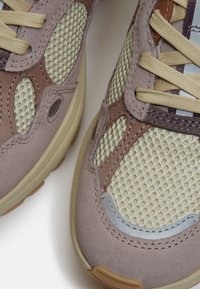 Gros plan sur des baskets beige et marron montrant un tissu en maille, des panneaux en daim et des lacets beiges sur un fond clair.