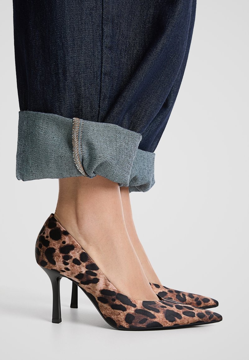 Vue des chevilles d'une personne portant un jean bleu foncé retroussé et des chaussures à talons hauts à bout pointu imprimées léopard marron sur un fond gris.