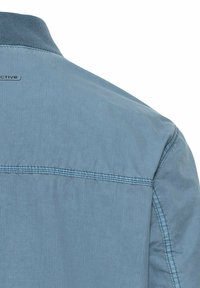 Lichtblauwe shirt met een gladde textuur, voorzien van een eenvoudige kraag en horizontale stiknaden over de rug. Subtiele branding bij de halslijn.
