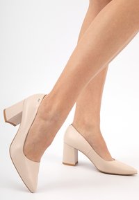 Scarpe con tacco alto beige con una finitura in pelle liscia, punta affusolata e tacco quadrato largo, caratterizzate da un design minimalista e linee pulite.